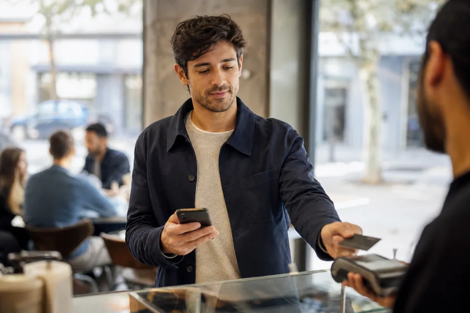 Man paying with a metal credit card at a cafe while checking his balance on his phone