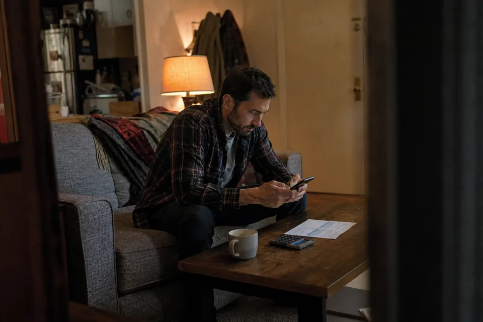 Renter on a couch checking the monthly fees on his rent payment app next to a printed rent statement