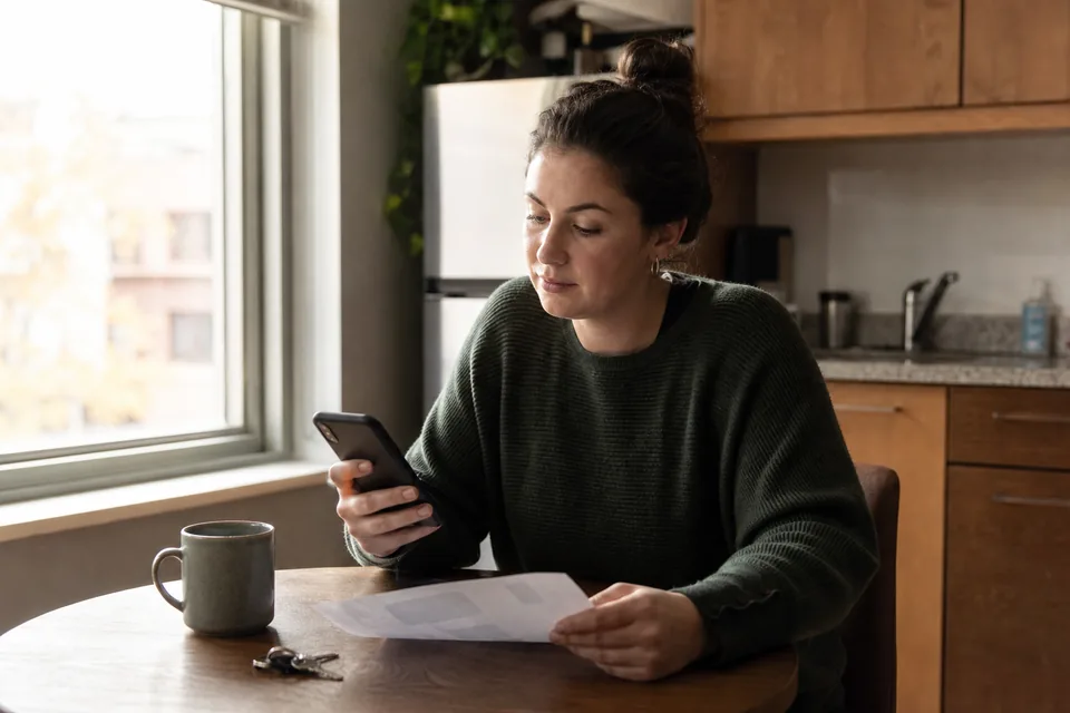 Renter at her kitchen table checking a rent payment on her phone next to a printed bill
